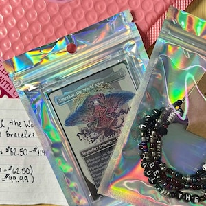 May include: Two sealed holographic bags containing a trading card and a beaded bracelet. The card features the text "Emrakul, the World Anew". The bracelet has black beads with white letter beads spelling "THE WORLD ANEW". A pink bubble mailer is in the background.