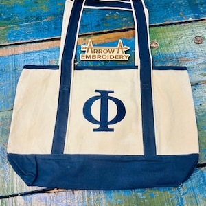 Könnte beinhalten: Cremefarbene Tragetasche mit marineblauen Akzenten und einem großen marineblauen griechischen Buchstaben Phi. Die Tasche hat marineblaue Griffe und Besätze. Das Holzlogo von "ARROW EMBROIDERY" ist oben an der Tasche befestigt.