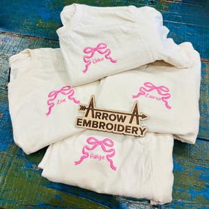 May include: Cream-coloured t-shirts featuring pink embroidered bows and names: Chloe, Zoe, Zaria, and Paige. A wooden sign with the text "ARROW EMBROIDERY" is also present. The shirts are displayed on a blue and green wooden surface.