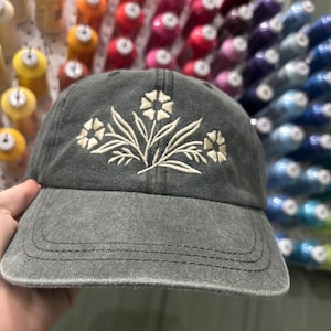 May include: A gray baseball cap with a floral embroidery design in cream-colored thread. The cap has a curved brim and a distressed look. The background shows spools of colorful thread.