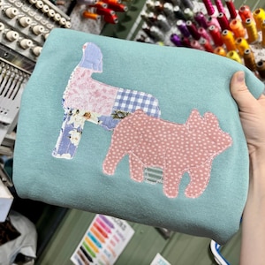 May include: A light blue sweatshirt featuring two fabric applique animal shapes. One shape is a standing dog made of floral and gingham patterned fabric, and the other is a pig shape made of pink fabric with a white polka dot pattern.