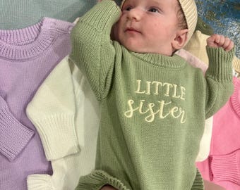 May include: A sage green knitted long-sleeve romper with the text LITTLE sister embroidered in white on the chest, featuring a ruffled hem and a matching beige ribbed knit headband with a large bow, surrounded by other colorful knit rompers.