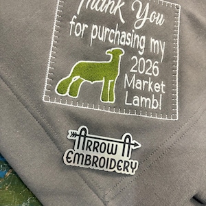 May include: A grey blanket featuring white embroidered text: "Thank You for purchasing my 2026 Market Lamb!" A green lamb graphic is also embroidered. A silver pin with "Arrow Embroidery" is attached to the blanket.