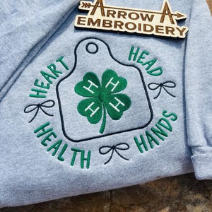 Puede incluir: Sudadera gris claro con bordados verdes. El diseño incluye un trébol de cuatro hojas y las palabras "HEART, HEAD, HEALTH, HANDS". Un letrero de madera con el texto "ARROW A EMBROIDERY" está encima.