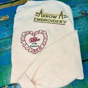 Könnte beinhalten: Ein hellrosa Sweatshirt mit einem herzförmigen gestickten Design mit der Aufschrift "Dr. Pepper is my Valentine". Das Sweatshirt hat auch ein pfeilförmiges Holzlogo mit der Aufschrift "Arrow Embroidery".