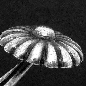 May include: A silver daisy-shaped button with a raised center. The button has a textured surface and is designed to be sewn onto fabric.