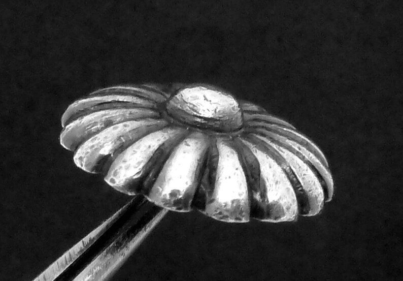 May include: A silver daisy-shaped button with a raised center. The button has a textured surface and is designed to be sewn onto fabric.