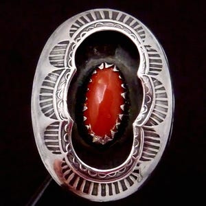 May include: Oval-shaped silver pendant with a vibrant red stone center. The pendant features intricate stamped designs around the edge, creating a detailed and textured appearance. The red stone is oval-shaped and surrounded by a silver setting.