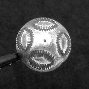 May include: A small, round, silver-toned metal bead featuring a textured surface with four distinct, oval-shaped stamped patterns arranged symmetrically around a central point. The bead has a metallic finish and a small hole in the centre.