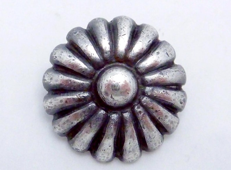 May include: A silver-toned metal button with a raised, circular center and a decorative, sunburst-like design around the edge.
