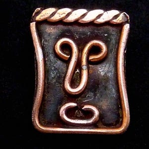 May include: A rectangular copper-toned metal pendant with a dark, textured centre. The pendant features a stylized design with a central figure resembling a face, and a braided border. The design is likely inspired by ancient or tribal art.