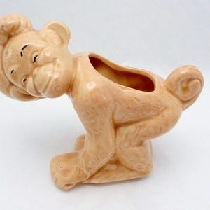 Pode incluir: Um vaso de cerâmica em forma de macaco, com um esmalte marrom claro. O macaco está sentado sobre os calcanhares com a cabeça inclinada para trás e os braços estendidos. O vaso tem uma pequena abertura na parte de trás para plantar.