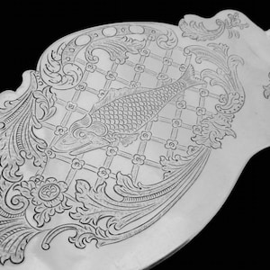 May include: Close-up of a silver serving utensil with intricate engraved details. The design features a fish within a grid pattern, surrounded by floral and scrollwork motifs. The handle extends from the decorative head.