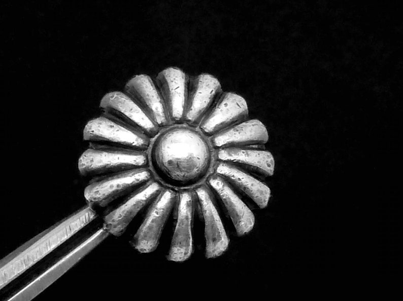 May include: A silver-toned metal flower-shaped decorative piece with a raised center. The flower has a textured surface and a circular center.