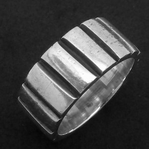 Puede incluir: Un anillo plateado con una banda ancha. El anillo presenta una serie de ranuras rectangulares verticales que rodean la banda. El diseño le da al anillo una apariencia texturizada. El anillo se muestra sobre un fondo oscuro.