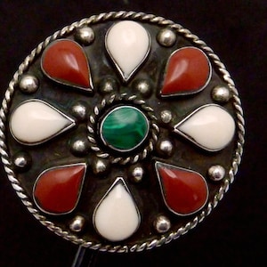 May include: A round, silver-toned brooch with a central green stone, surrounded by alternating red and white teardrop-shaped stones. The brooch has a twisted rope border and small silver accents.