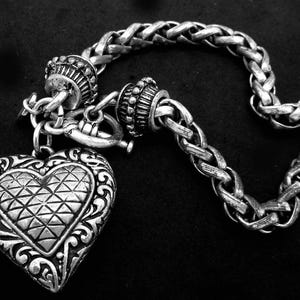 May include: A silver-toned bracelet with a braided chain design and a heart-shaped pendant. The heart pendant features an intricate geometric pattern and decorative scrollwork. The bracelet is displayed against a black background.