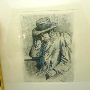 May include: Framed etching of a man in a hat, leaning on a table with a cane. The artwork is in black and white, with fine lines creating the image. The frame is gold with a cream-colored matting.