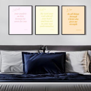 May include: Three framed prints with inspirational quotes in a bedroom setting. The prints feature text in various colors against pastel backgrounds. The bed has gray and white bedding with navy blue pillows and a blanket. Black lamps and a side table are on either side.