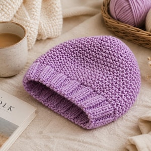Peut inclure: Un bonnet en tricot lilas avec un bord côtelé. Le chapeau est fait de fil doux et est présenté sur une surface de couleur neutre. L'image comprend également une tasse de café, un livre et des pelotes de laine dans un panier.