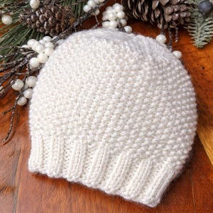 May include: A cream-coloured knitted beanie hat. The hat has a ribbed brim and a textured stitch pattern on the crown. The hat is displayed on a wooden surface with decorative pine cones and greenery in the background.