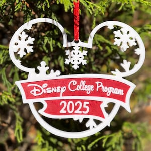 May include: White and red Christmas ornament in the shape of Mickey Mouse ears with snowflake accents. The ornament has a red banner that reads "Disney College Program 2025". A red ribbon is attached for hanging on a Christmas tree.