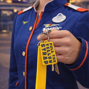 May include: A blue uniform with orange trim and gold buttons. The uniform has a "Soarin" logo and a name tag. A yellow key tag with the text "Pull the Yellow Strap" is attached to a set of keys.