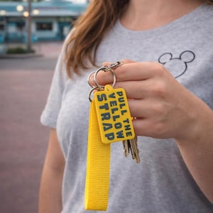 May include: A yellow keychain with the text "PULL THE YELLOW STRAP" in blue. The keychain has a matching yellow strap and is attached to a set of keys. The person in the image is wearing a gray t-shirt.