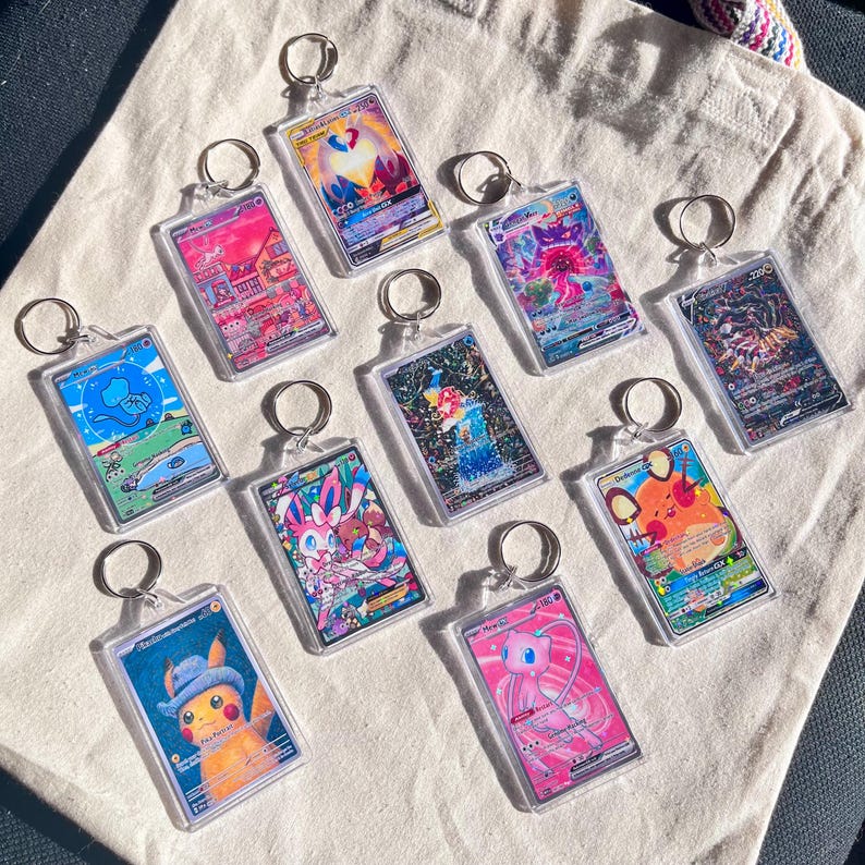 May include: A collection of clear acrylic keychains featuring colorful illustrations of various characters. Each keychain has a silver metal ring. The illustrations showcase a variety of designs, including a Pikachu wearing a hat and a pink Mew.