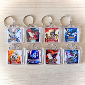 Puede incluir: Una colección de ocho llaveros de acrílico transparente con varias portadas de juegos de Pokémon. Cada llavero tiene un anillo de metal plateado y muestra arte de juego vibrante, incluyendo títulos como Pokemon X, Omega Ruby y Ultra Moon. Los llaveros son rectangulares y parecen nuevos.