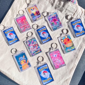 May include: A collection of clear acrylic keychains featuring various Pok&eacute;mon trading card designs. Each keychain has a silver ring and showcases colorful illustrations of Pok&eacute;mon characters. The backs of the keychains display the Pok&eacute;mon logo.