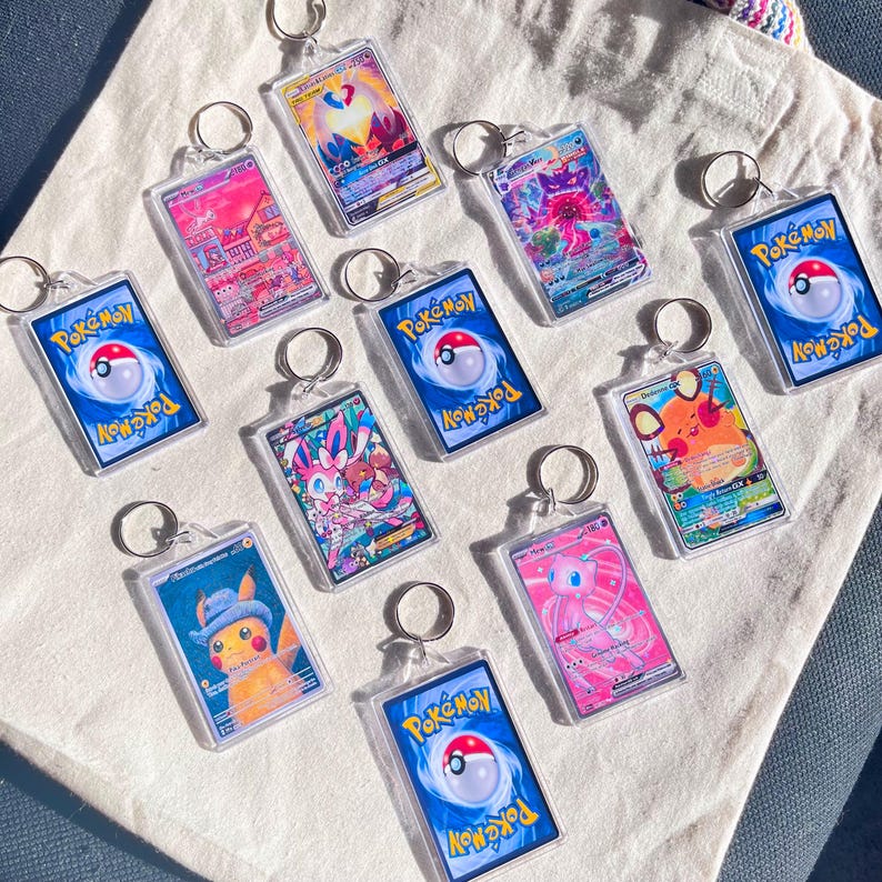 May include: A collection of clear acrylic keychains featuring various Pok&eacute;mon trading card designs. Each keychain has a silver ring and showcases colorful illustrations of Pok&eacute;mon characters. The backs of the keychains display the Pok&eacute;mon logo.