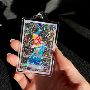 May include: A clear rectangular keychain featuring a holographic trading card with a colorful illustration of a fish-like creature and the text "Magikarp". The card is encased in a clear plastic holder.