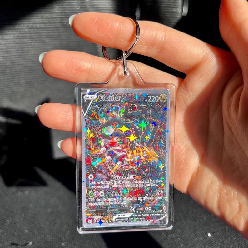 May include: A Giratina V Pokemon card keychain. The card features a holographic design with a colorful illustration of Giratina. The keychain has a silver metal ring and a clear plastic case.