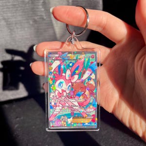 May include: A clear acrylic keychain featuring a Sylveon EX Pokemon trading card. The card has a colorful design with pink, blue, and brown accents. The keychain includes a silver metal keyring.