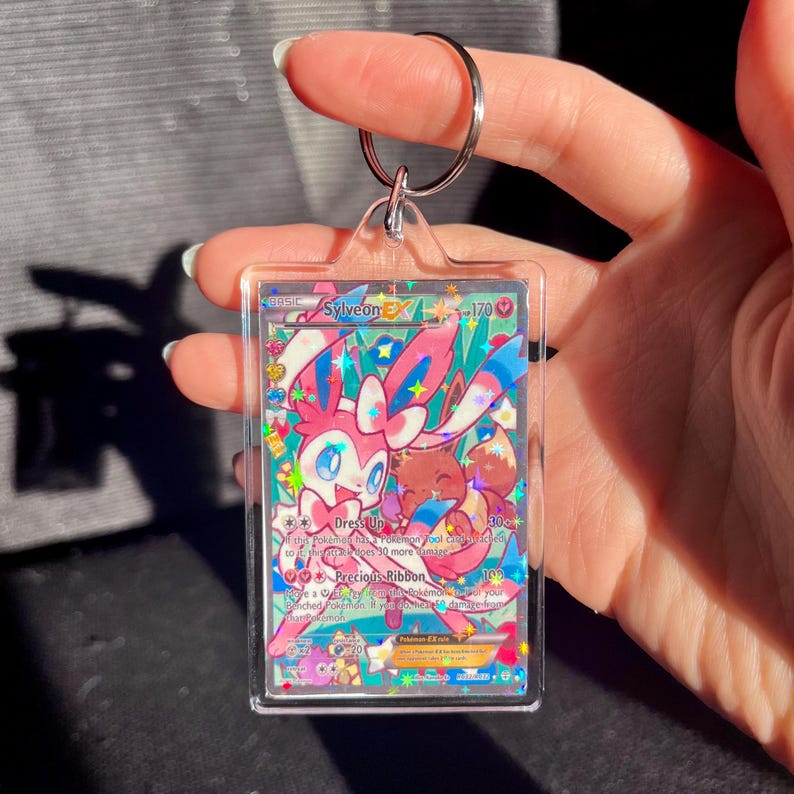 May include: A clear acrylic keychain featuring a Sylveon EX Pokemon trading card. The card has a colorful design with pink, blue, and brown accents. The keychain includes a silver metal keyring.