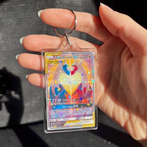 May include: A clear acrylic keychain featuring a Pok&eacute;mon trading card. The card displays Latias & Latios GX with a colorful design of blue, red, and yellow hues. The card is encased in a protective sleeve and attached to a silver keyring.