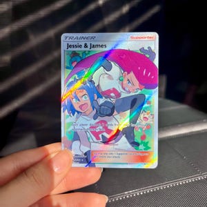 May include: A holographic Pokémon trading card featuring Jessie and James. The card displays a colourful illustration of the characters, with the text "Jessie & James" and "Supporter". The card is held in a protective sleeve.