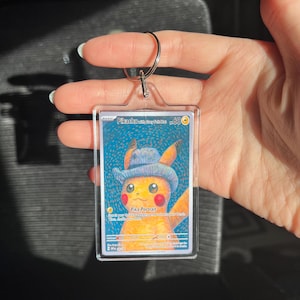 May include: A clear acrylic keychain featuring a Pikachu card with a cowboy hat. The card has a blue background and the text "Pikachu with Cowboy Hat". The keychain has a silver metal ring.