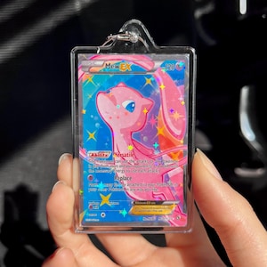 May include: A pink Mew EX Pok&eacute;mon trading card in a clear acrylic keychain. The card features a pink Mew character with blue eyes and a blue and pink background. The card has text that reads "Ability Versatile" and "Replace".