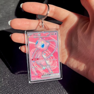 May include: A pink and white Mew EX Pokemon trading card encased in a clear acrylic holder with a silver keyring. The card features a pink Mew character with blue eyes and the text "Mew EX".