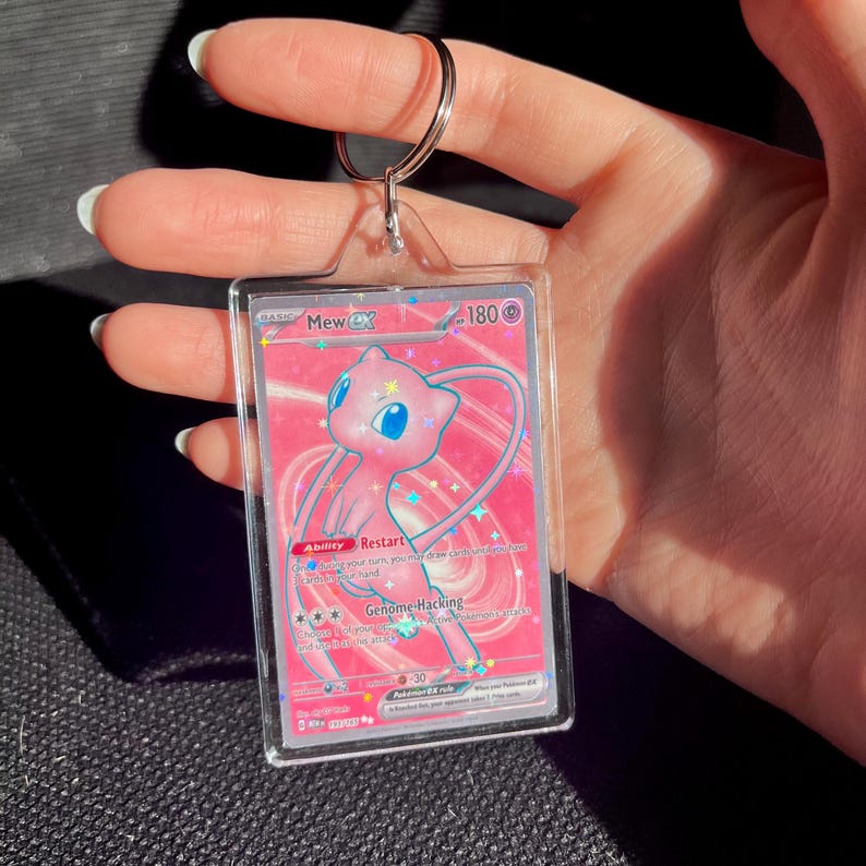 May include: A pink and white Mew EX Pokemon trading card encased in a clear acrylic holder with a silver keyring. The card features a pink Mew character with blue eyes and the text "Mew EX".