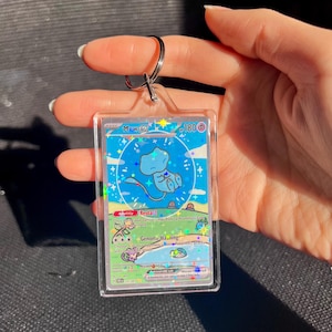 May include: A clear acrylic keychain featuring a Pok&eacute;mon trading card with a blue Mew ex character. The card has a holographic design with text and a silver keyring attached at the top.