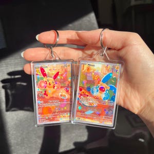 May include: Two clear acrylic keychains featuring colorful illustrations of cartoon characters. Each keychain has a silver metal ring. The cards have text and are held by a hand.