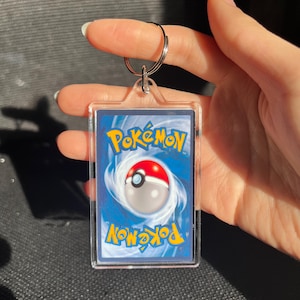 May include: A clear acrylic keychain featuring a Pok&eacute;mon trading card. The card has a blue background with the Pok&eacute;mon logo in yellow and a red and white Pok&eacute; Ball design. A silver keyring is attached at the top.