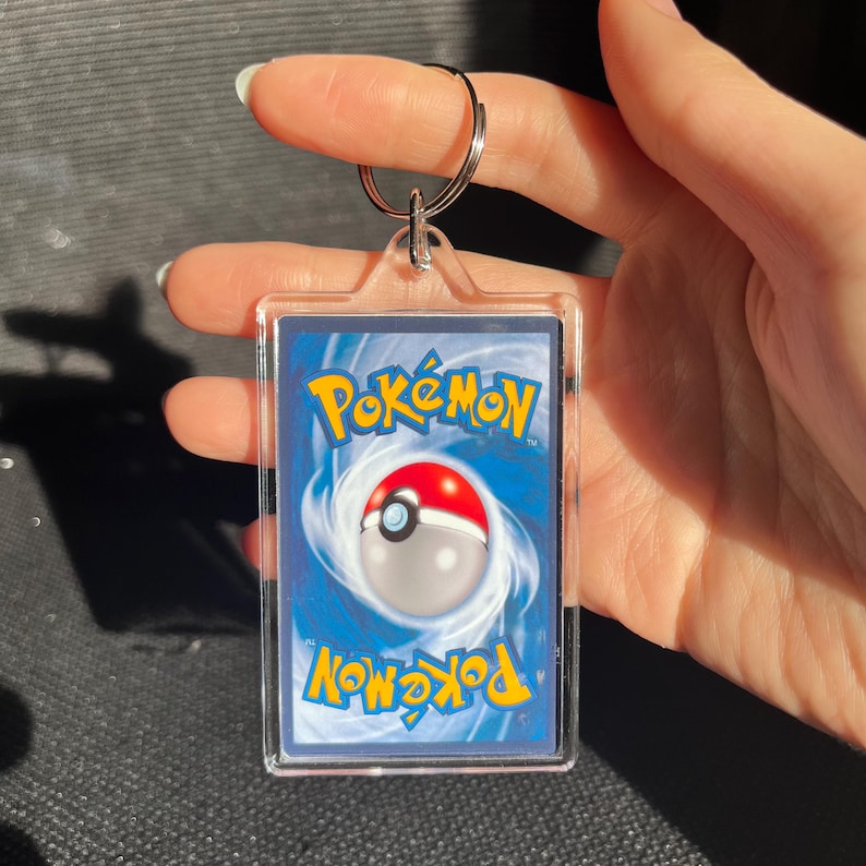 May include: A clear acrylic keychain featuring a Pok&eacute;mon trading card. The card has a blue background with the Pok&eacute;mon logo in yellow and a red and white Pok&eacute; Ball design. A silver keyring is attached at the top.