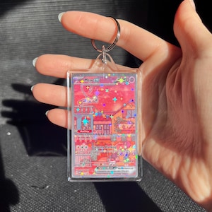 May include: A clear acrylic keychain featuring a holographic Pokemon card with a pink and red background. The card has the text "Mew EX" and other details. A silver keyring is attached to the top.