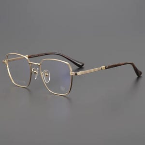 Puede incluir: Gafas con montura dorada de forma cuadrada y un sutil emblema floral en la lente. Las patillas son de veta de madera oscura, y las monturas tienen un dise&ntilde;o de doble barra. Las gafas est&aacute;n sobre un fondo gris.