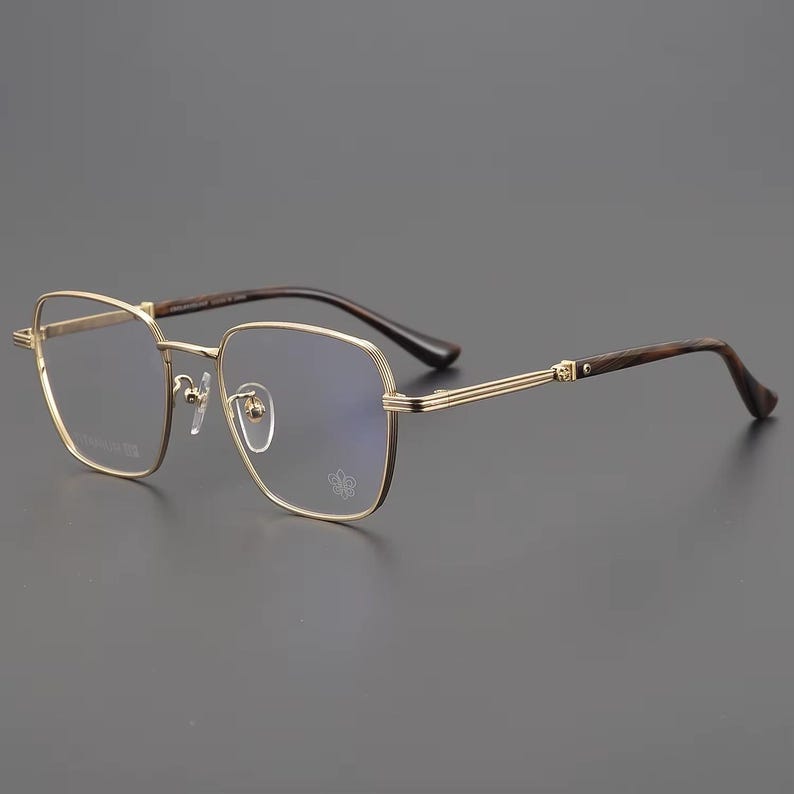 Puede incluir: Gafas con montura dorada de forma cuadrada y un sutil emblema floral en la lente. Las patillas son de veta de madera oscura, y las monturas tienen un dise&ntilde;o de doble barra. Las gafas est&aacute;n sobre un fondo gris.