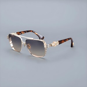 May include: Gold-coloured sunglasses with gradient lenses. The sunglasses have a double bridge and decorative detailing on the temples. The arms feature a tortoiseshell pattern. The lenses transition from light to dark.